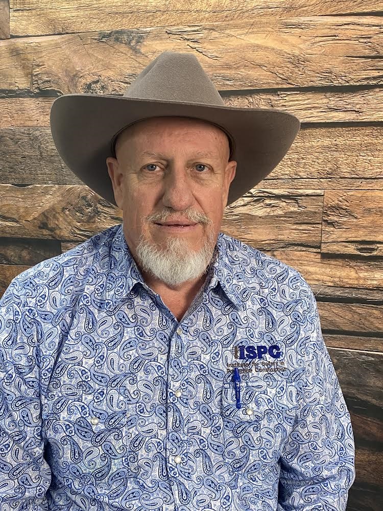 A white man with a blue goatee, wearing a blue, collared paisley shirt with the ISPC logo and a brown cowboy hat looks at the camera. Mack Turner