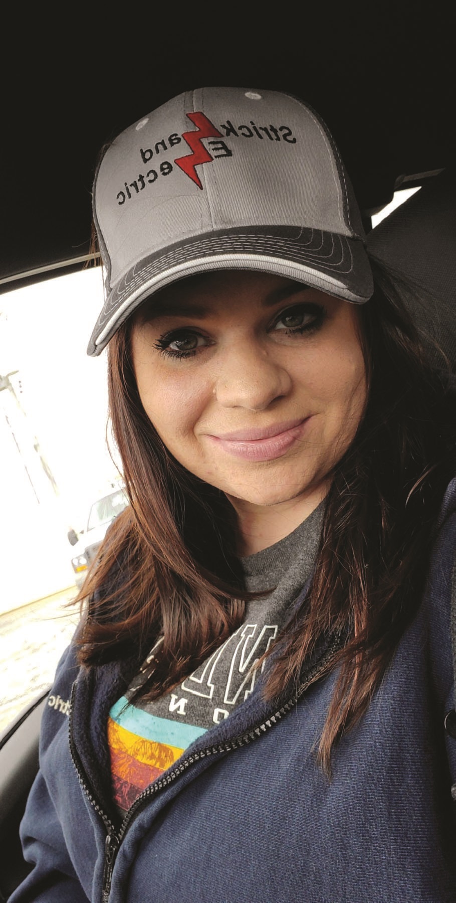 A woman, Abbey Strickland, wears a Strickland Electric hat and smiles in a car. Image by Abbey Strickland.