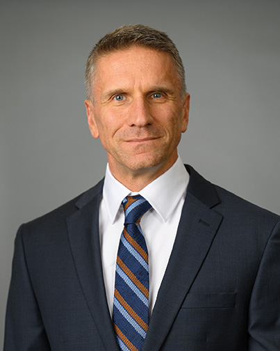 A man, Edmund Milukas, smiles at the camera in front of a gray background. 