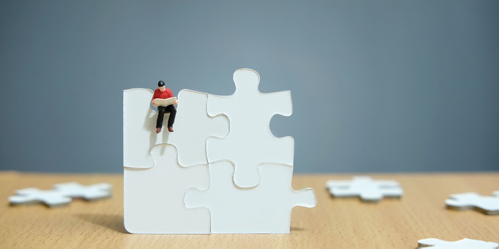 Four connected puzzle pieces stand upright, surrounded by other scattered puzzle pieces on the table. A man sits on the upright puzzle pieces looking at a book.