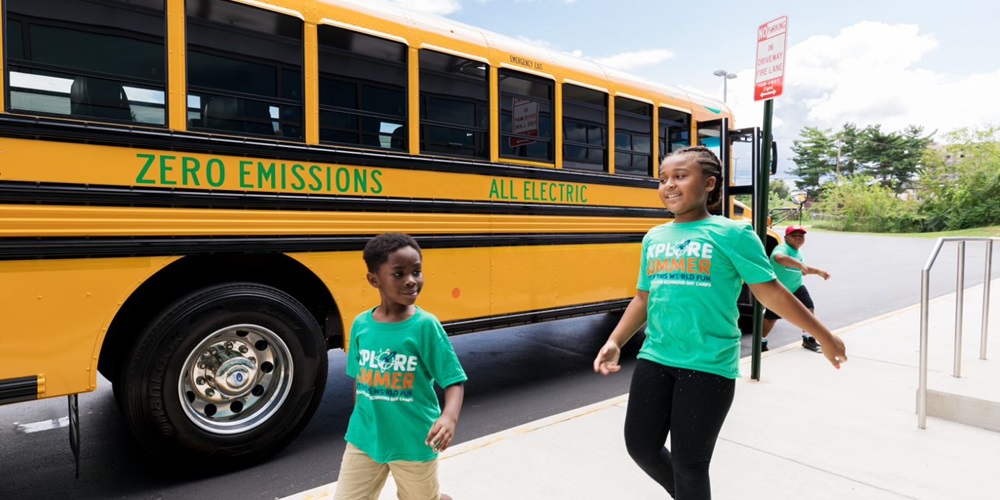 Two children walk past a school bus which reads Zero Emissions all electric along the side.
