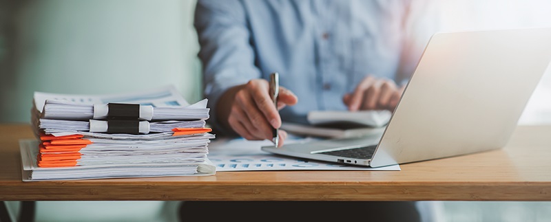 A person sits at a desk in front of a computer and a pile of papers, representing someone keeping project documentation organized.