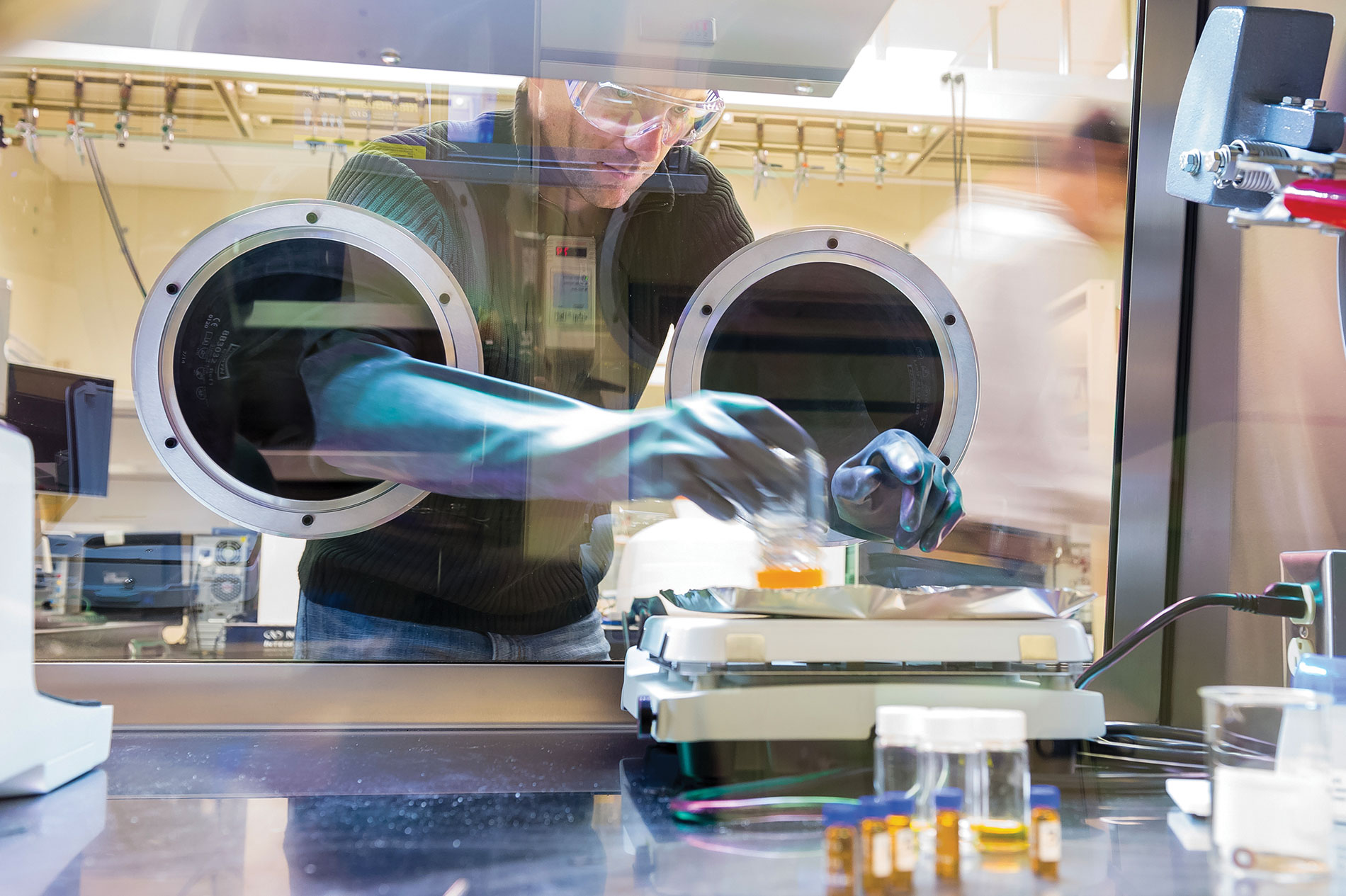 A man with gloves reaches into a sealed glass area with vials. Image by Argonne National Laboratory.