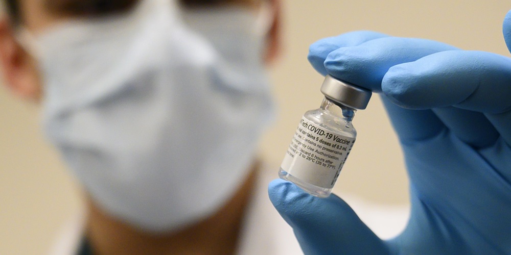 A man wearing a mask and gloves holding a vial of the Pfizer-BioNTech COVID-19 vaccine
