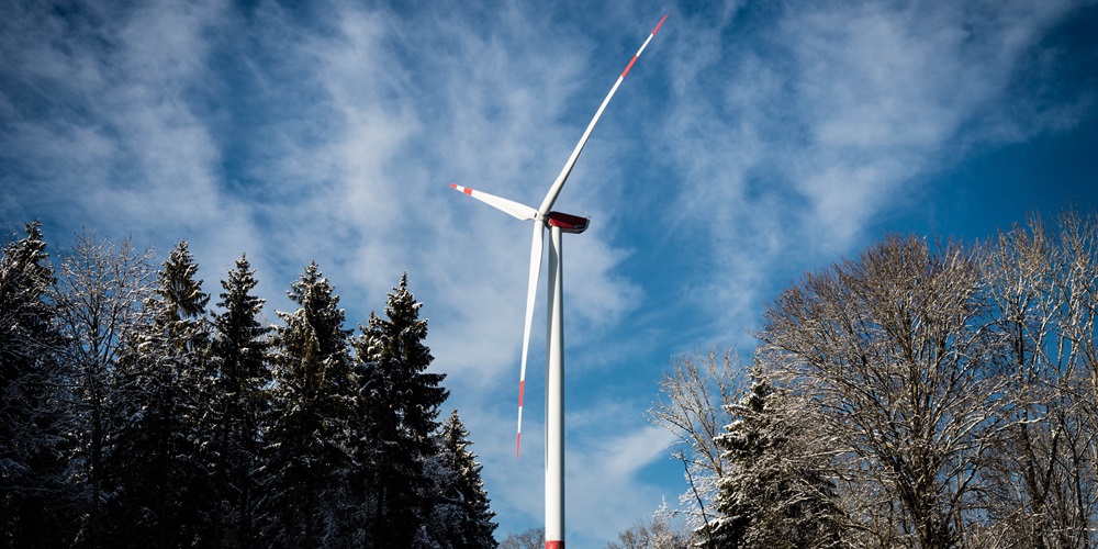 A wind turbine framed by trees set against a blue sky with wispy clouds