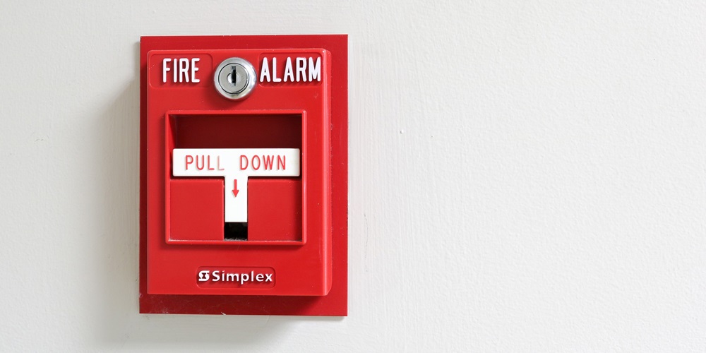 Red manual fire alarm pull station mounted on a wall, representing a key initiating device in both conventional and addressable fire alarm control systems.