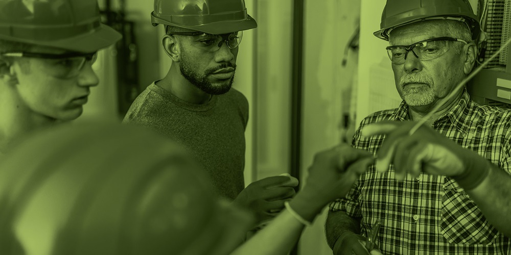 An older man in a plaid shirt and hardhat demonstrates a skill to several other people standing around