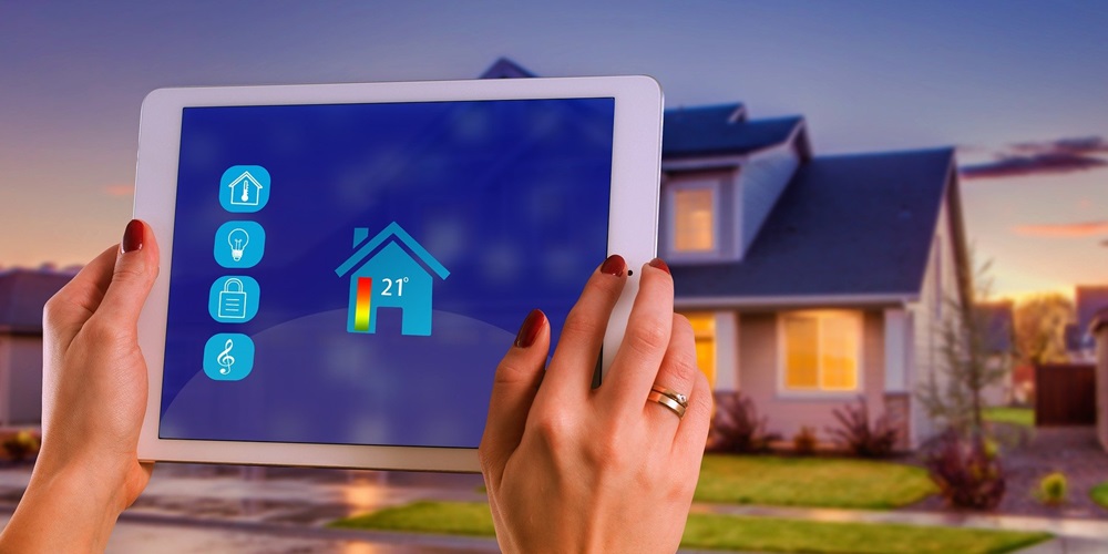 A person's hands holding a tablet showing a smart home heating system, with the home in the background
