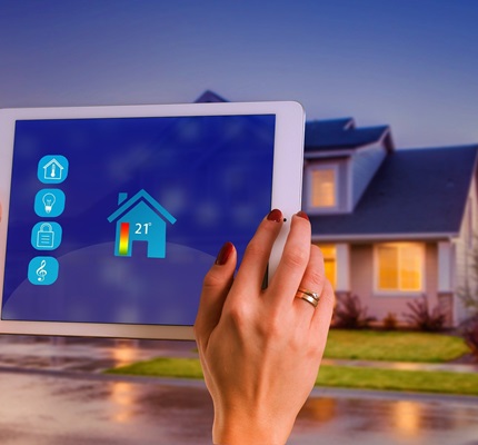 A person's hands holding a tablet showing a smart home heating system, with the home in the background