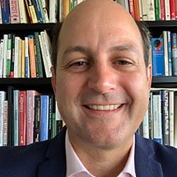 A man, Andrew McCoy, smiles in front of a bookshelf. Image by Andrew McCoy.