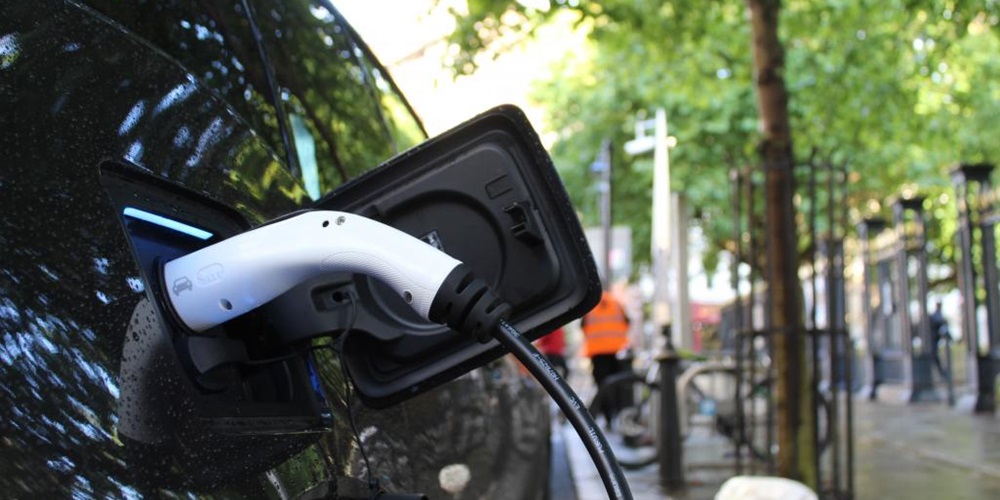 A car charges with an EV charger on a tree-lined street. Image by Unsplash / Andrew Roberts.