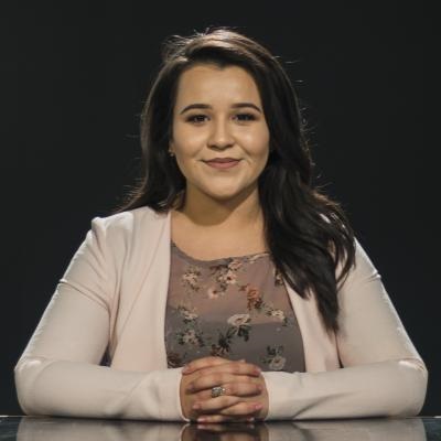 Annabel Rocha A woman in a flowered shirt and white cardigan, with brown hair, sits at a table against a black background