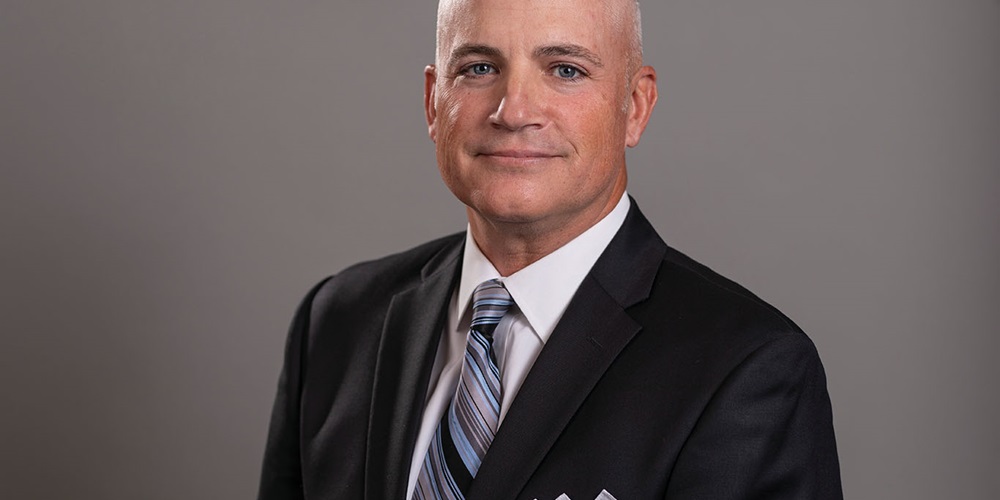 A man in a suit, O’Brien Mills, smiles in front of a gray background. Image by O’Brien Mills.