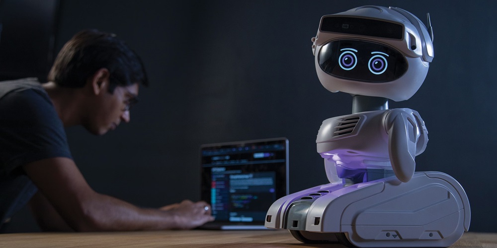 A small, cute robot sits on a table while a man hunches over a computer in the background | Misty Robotics