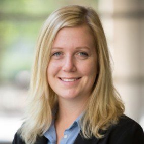 A woman with blonde hair wearing a blue collared shirt and black sweater smiles at the camera