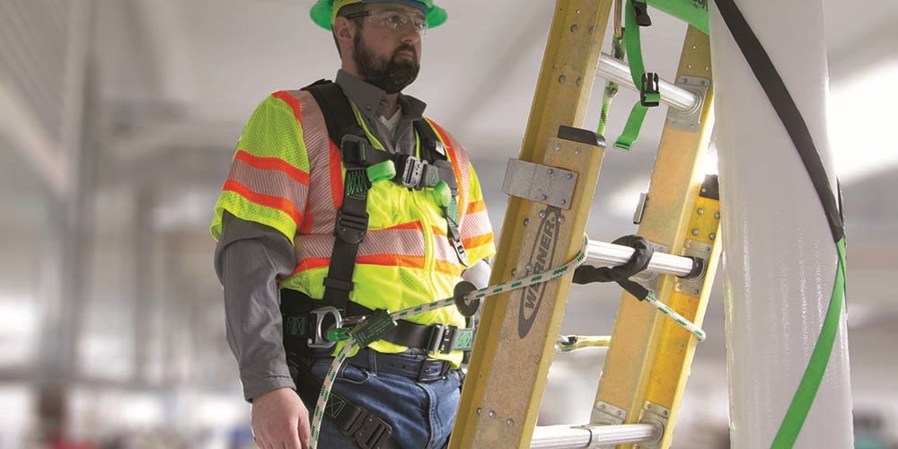 A worker in a green Buckingham helmet and harness stands on a ladder indoors. Image by Buckingham Manufacturing.