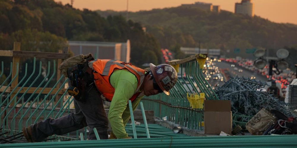 A worker moves rebar on a rooftop against a sunset hill. Image by Chandler Denise for Unsplash.