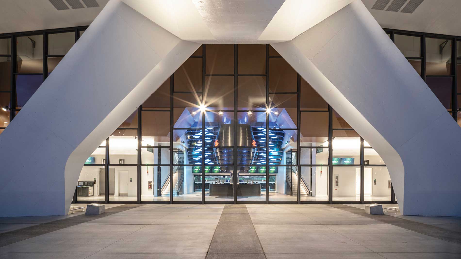 A colorful, lit-up arena entry hall framed by angled concrete pillars outside. Image by Alex Fradkin Photography.