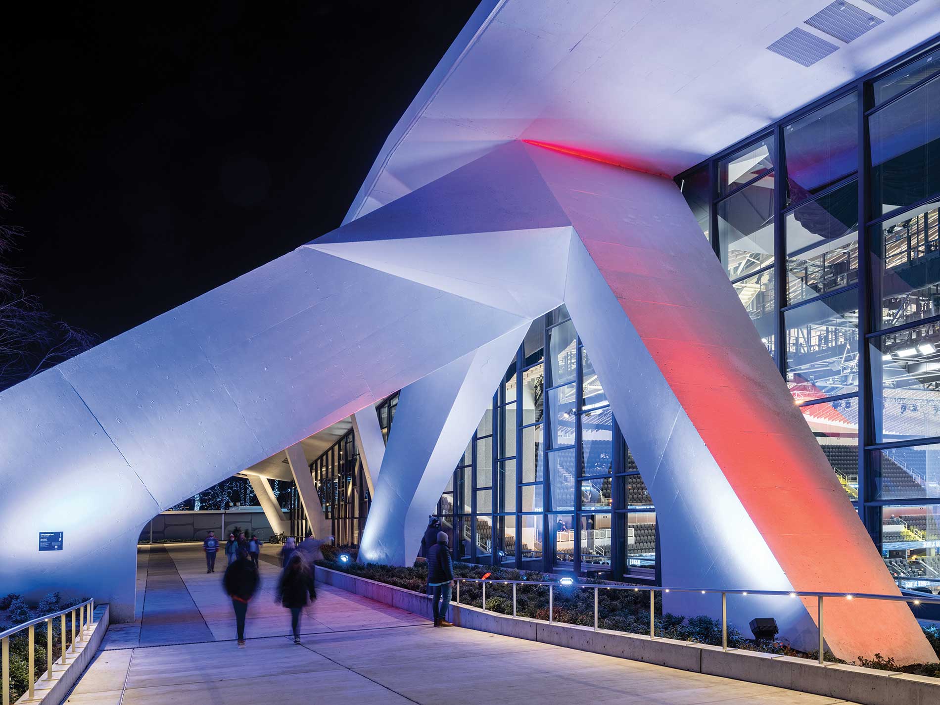 A sculptural, concrete exterior wall and concrete walkway are colorfully lit up. Image by Alex Fradkin Photography.