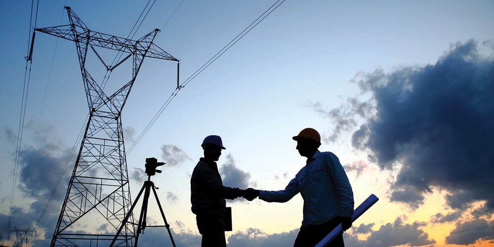 Two people in hard hats shake hands, silhouetted against a transmission tower, tripod and sunset. Image by Getty Images / Aydin Mutlu.