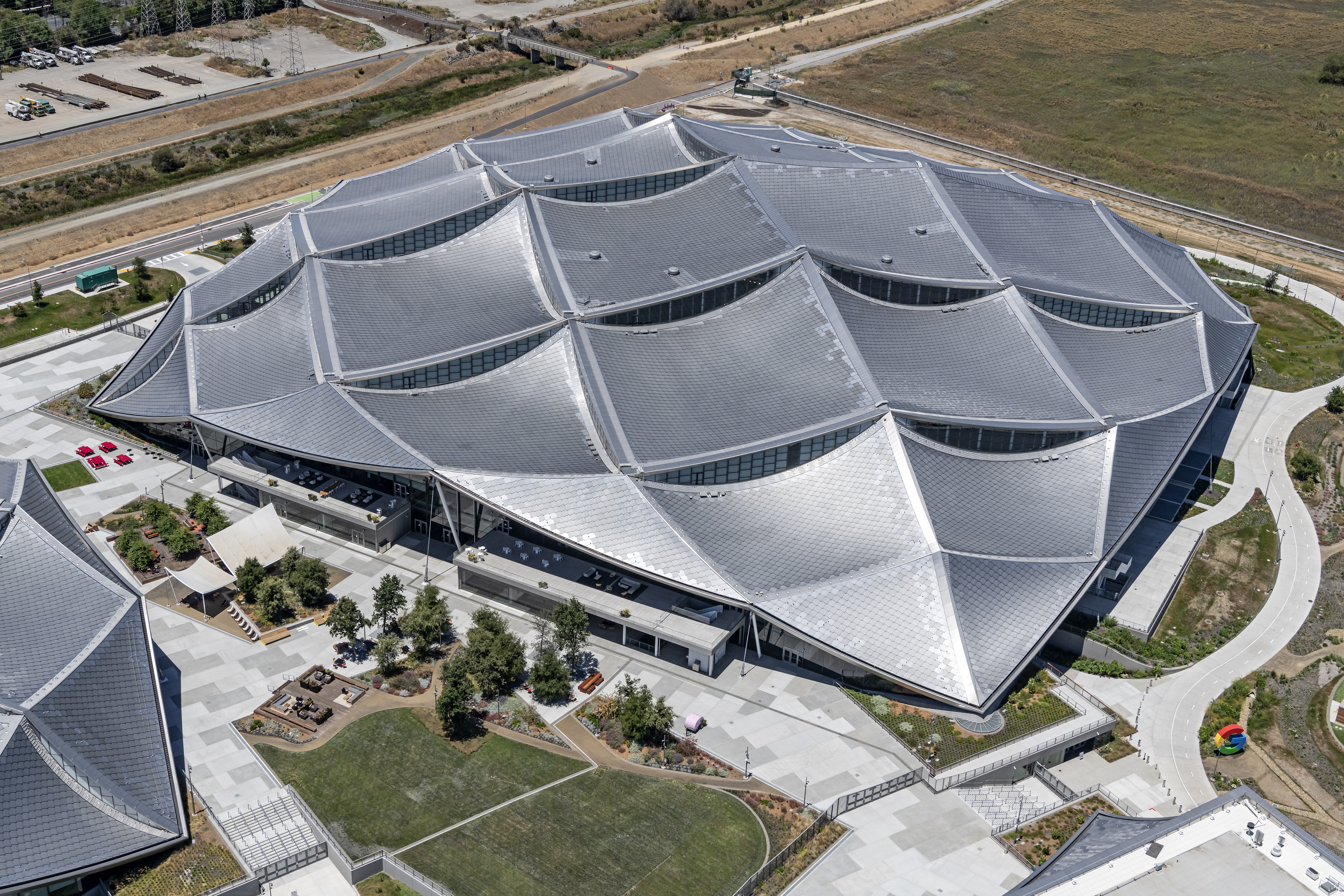 A large building surrounded by grass and concrete, with sail-shaped roof panels covered in gray solar panels. 