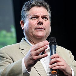 A man, Gordon Feller, holds a microphone in front of a black and green background. Image by Gordon Feller.