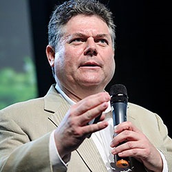 A man, Gordon Feller, holds a microphone in front of a black and green background. Image by Gordon Feller.
