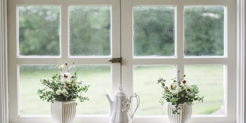Three white vases with greenery in front of a white window and lawn.