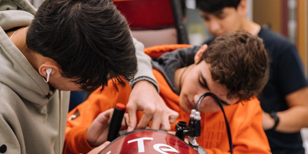 Students lean over a metal machine indoors. Image by Jeswin Thomas / Unsplash.