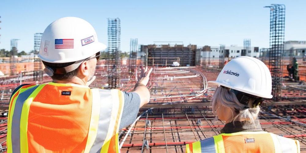 Two workers in vests and hard hats look over a roof. Image by Unsplash / Joe Holland.