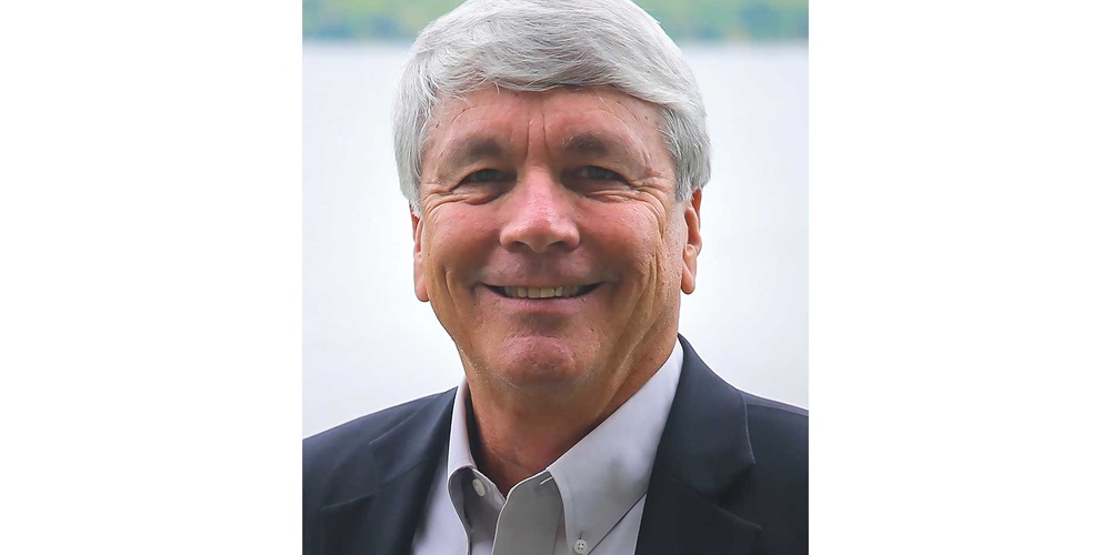 A man, Kirk Davis, smiles in front of trees and water.