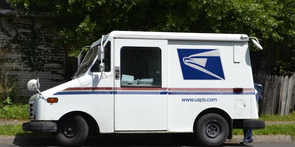 White USPS truck in front of a wall, fence and trees.