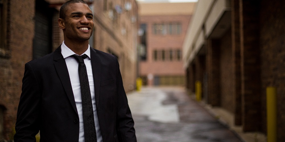 A black man in a suit and tie stands in an empty road and smiles. | Taylor Grote / Unsplash
