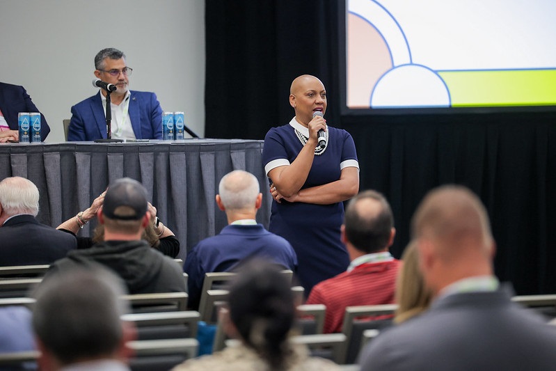 A speaker holds a microphone and presents to an audience at NECA 2024 San Diego