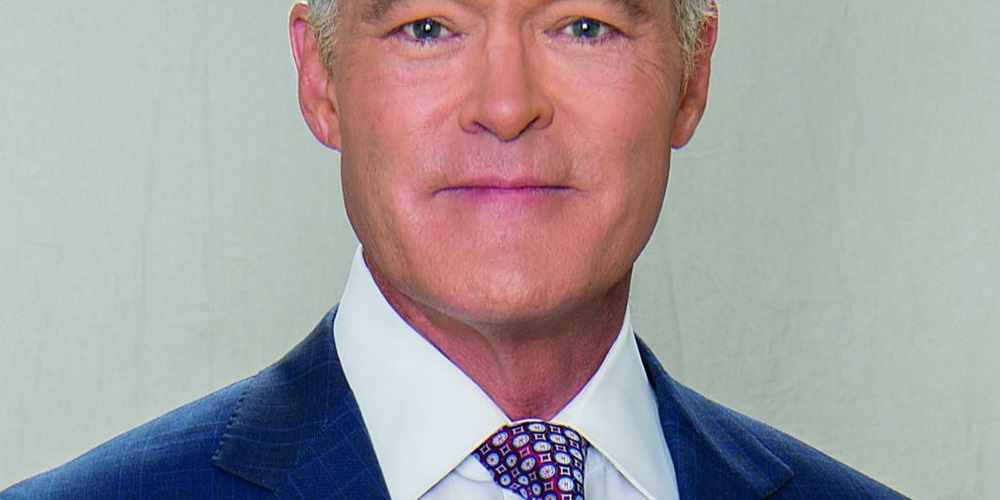 A man with white hair wearing a blue suit jacket, white shirt, and purple patterned tie