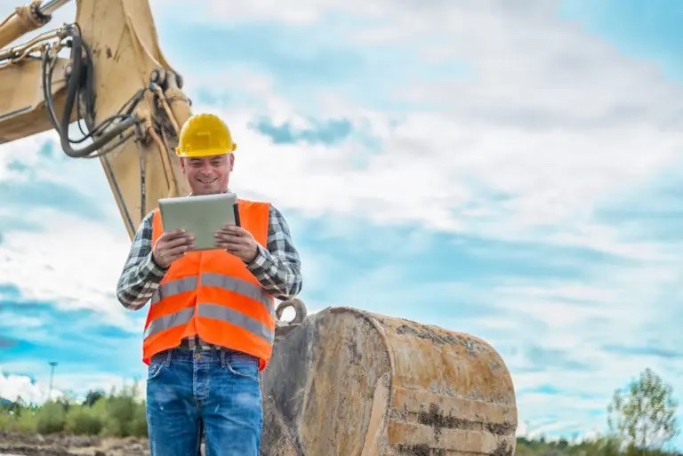 A man in jeans, a checked shirt, and hi-vis vest looks at a clipboard, standing in front of a backhoe
