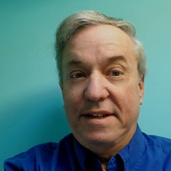 A man, Richard Bingham, smiles in front of a blue background. Image by Richard Bingham.