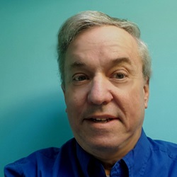 A man, Richard Bingham, smiles in front of a blue background. Image by Richard Bingham.