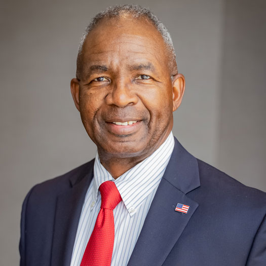Ronald Bailey headshot. A black man with grey hair in a blue suit, striped shirt and red tie smiles at the camera.