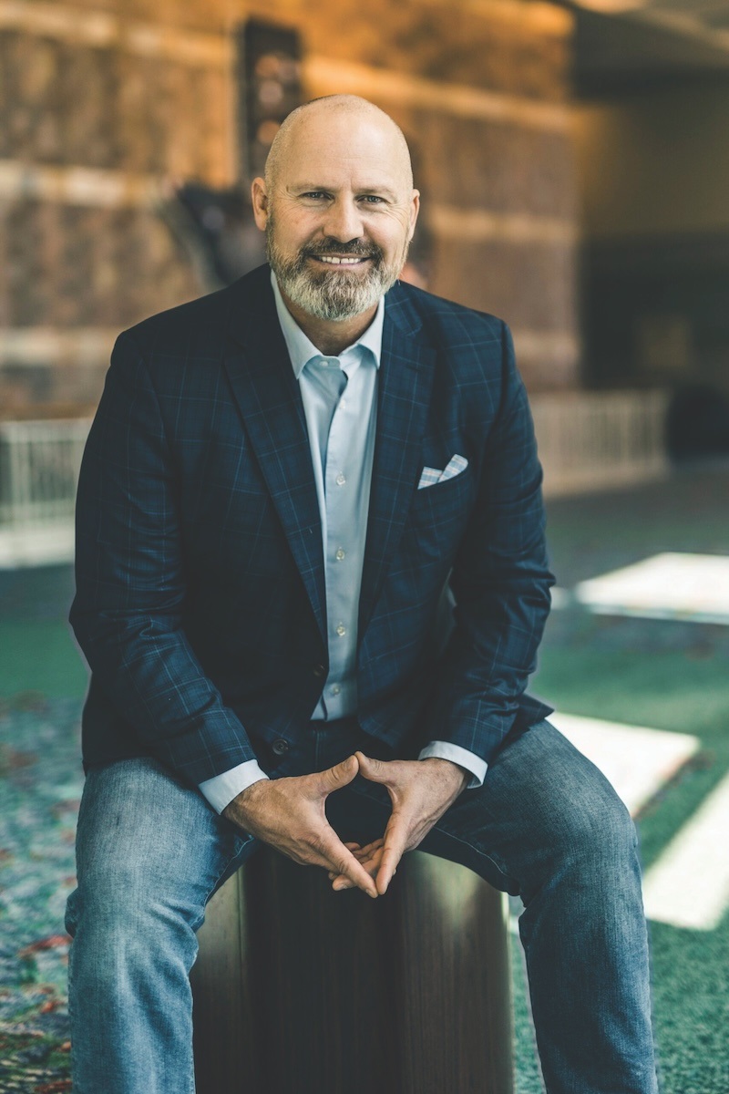 Rorke Denver. A balding man wearing jeans, a light blue collared shirt, and a navy blue blazer sits on a stool