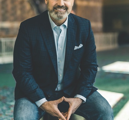 Rorke Denver. A balding man wearing jeans, a light blue collared shirt, and a navy blue blazer sits on a stool