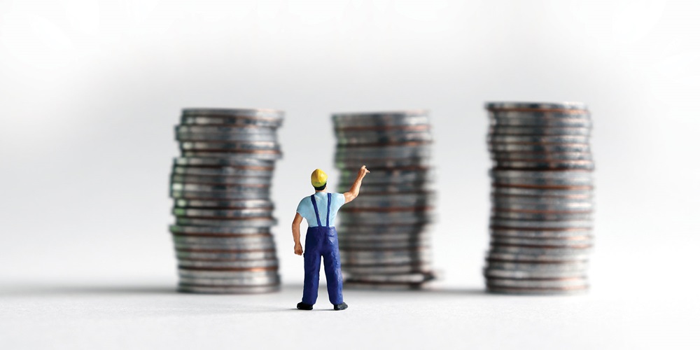 A toy model of a worker points up at three stacks of coins. Image by Shutterstock / Hyejin Kang.