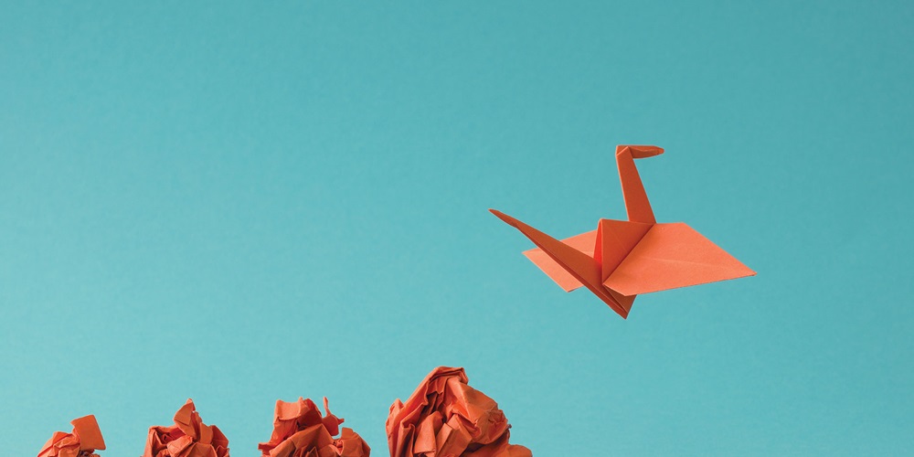 A row of four increasingly bigger pieces of crumpled orange paper against a blue background, with an orange origami crane in the air at the end. Image by Shutterstock / BeanImages.