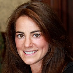 A woman, Susan Bloom, smiles in front of a tan background. Image by Susan Bloom.