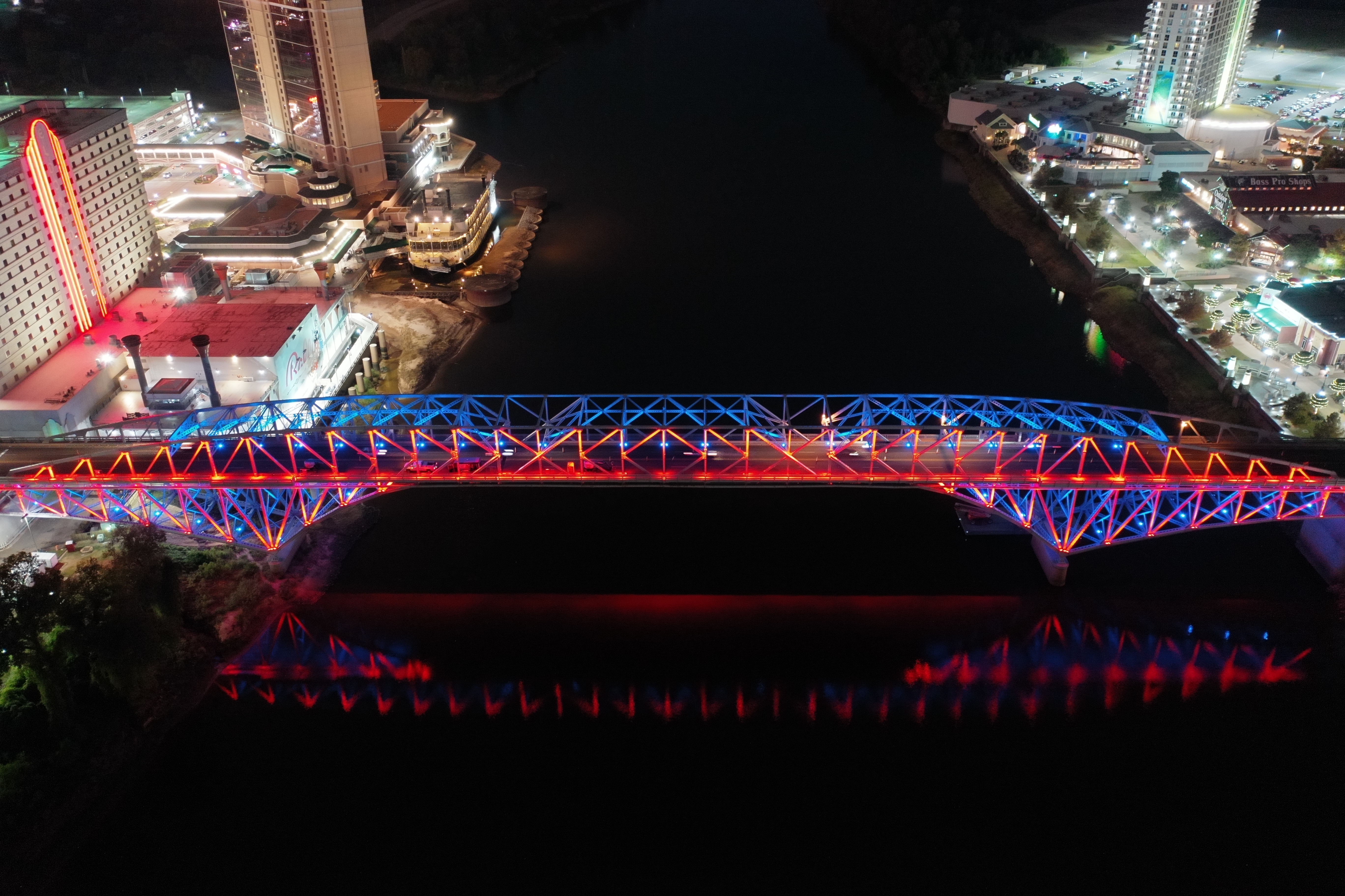 A bridge with blue and red lighting spans a dark body of water at night. Buildings line either end. 